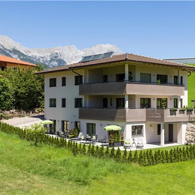 A modern residential house surrounded by green meadows and mountains. The sun is shining, and there is a clear blue sky.