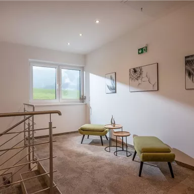 A bright staircase with two green armchairs and a small table. Decorative pictures hang on the wall, and a large window lets in plenty of daylight.