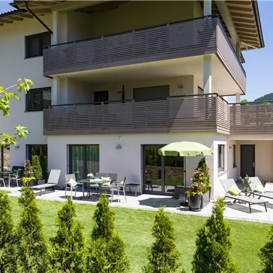 A modern building with a spacious balcony. The garden is green, equipped with loungers and a sun umbrella.