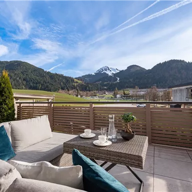 A cozy terrace with a seating area and a table. In the background, green hills and snow-capped mountains can be seen.
