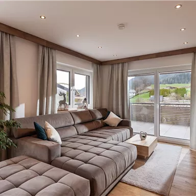 A modern living room with a comfortable gray sofa and large windows. In the background, a green landscape and gentle hills can be seen.