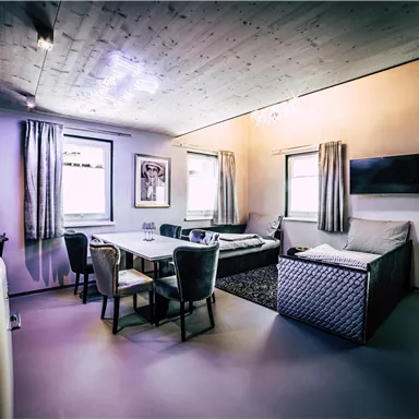 A modern living room with a dining table and comfortable chairs. There are windows with curtains and a TV on the wall.