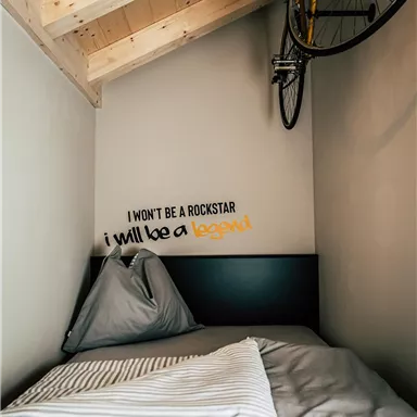 A small bedroom with a bed and a bicycle on the wall. On the wall, there is the saying: "I won't be a rockstar, I will be a legend."
