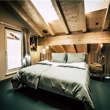 A cozy bedroom with slanted wooden beams and large windows. The bed is dressed with gray bedding, and the ambiance feels modern and inviting.