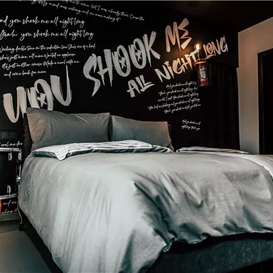 A modern bedroom with a large bed and a wall featuring artistic writings. Next to the bed is a drum, contributing to a creative ambiance.