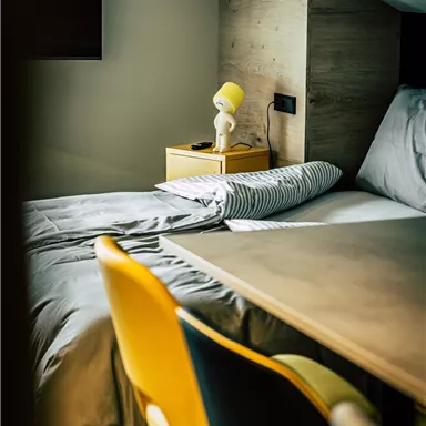A modern bedroom with a cozy bed and a small bedside table. Yellow chairs are next to a table that is well-lit.