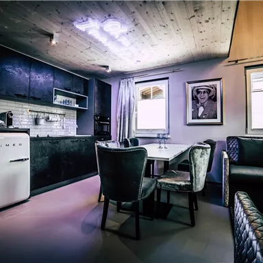 A modern kitchen with a dining table and stylish chairs. A picture hangs on the wall, and large windows let in plenty of light.