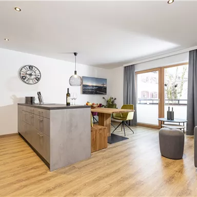 Ein modernes und helles Wohnzimmer mit offener Küche. Es gibt eine gemütliche Sitzecke und große Fenster, die viel Tageslicht hereinlassen.