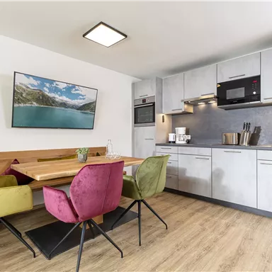 Moderne Küche mit Essbereich. Der Raum hat eine helle Holzoptik und farbige Stühle.