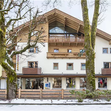 Ein modernes Gebäude mit Holzverkleidung und Balkonen, umgeben von schneebedeckten Bäumen. Es gibt eine klare Sicht auf die verschiedenen Etagen und Läden im Erdgeschoss.