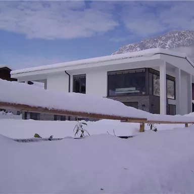 Ein modernes Haus im Schnee, umgeben von einer winterlichen Landschaft. Der Himmel ist klar und es liegen große Mengen frischer Schneedecke.