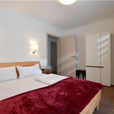 Ein modernes Schlafzimmer mit einem gemütlichen Bett und einem roten Überwurf. Die Wände sind in neutralen Farben gehalten, und es gibt einen Fernseher sowie einen Kühlschrank.