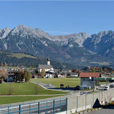 Eine malerische Landschaft mit hohen Bergen im Hintergrund und einem kleinen Dorf im Vordergrund. Die Umgebung ist grün und die Sonne scheint.