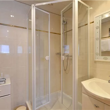 Ein modernes Badezimmer mit einer Dusche und einem Waschbecken. Die Wände sind hell gefliest und es gibt Handtücher auf einem Regal.