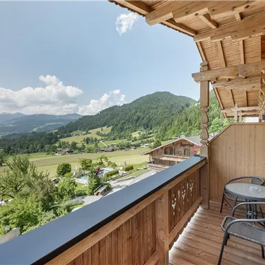 A balcony overlooking a green landscape and mountains. On the balcony are two chairs and a small table.