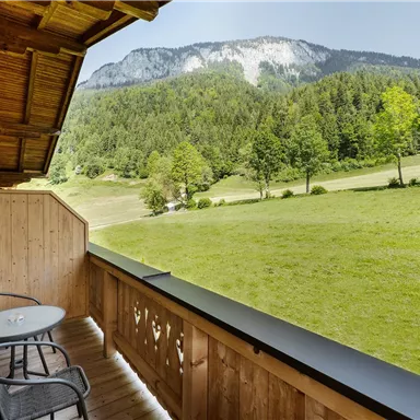A balcony with two chairs and a table that offers a view of a green meadow and mountains in the background. The surroundings are calm and surrounded by trees.