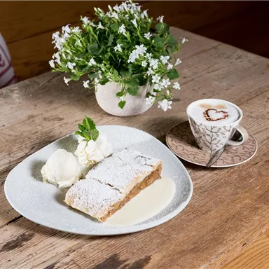 Ein Teller mit Apfelstrudel, Sahne und Vanillesauce liegt auf einem Holztisch. Daneben steht eine Tasse Kaffee mit einem Herzmotiv und ein kleiner Topf mit Blumen.