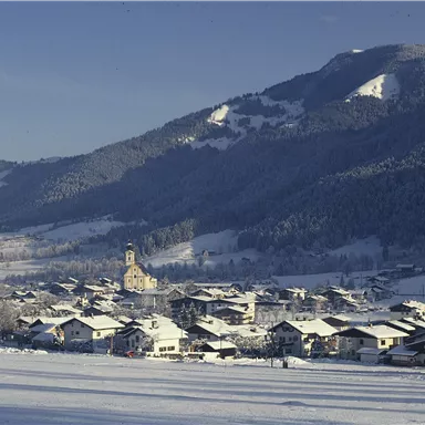 A snowy landscape with a small village and mountains in the background. The roofs of the houses are covered with snow and the surroundings are quiet and idyllic.
