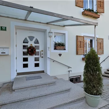 Ein einladender Hauseingang mit einer weißen Tür und schönen Fensterläden. Vor der Tür steht ein Blumentopf mit einem kleinen Baum.