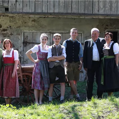 Eine Gruppe von sechs Personen in traditioneller Tracht steht vor einem alten Bauernhaus. Die Umgebung ist grün und sonnig.