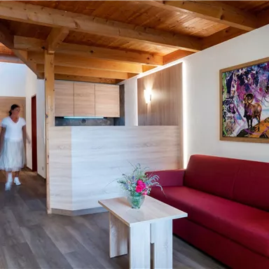 Ein modernes Wohnzimmer mit Holzdecke und rotem Sofa. Im Hintergrund ist eine Küchenzeile und ein farbenfrohes Gemälde zu sehen.