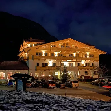 Ein gemütliches Hotel bei Nacht, beleuchtet von warmem Licht. Im Vordergrund sind einige Autos und schneebedeckter Boden zu sehen.