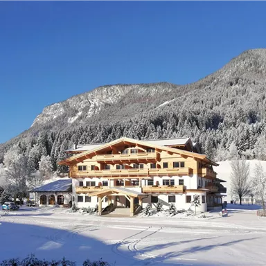 Ein gemütliches Hotel im Schnee, umgeben von verschneiten Bergen und Bäumen. Der klare blaue Himmel verleiht der Winterlandschaft eine besondere Schönheit.