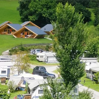 Eine grüne Landschaft mit modernen Holzhäusern im Hintergrund. Im Vordergrund sind Wohnmobile und Zelte auf einem Campingplatz zu sehen.
