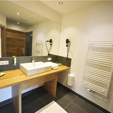 Ein modernes Badezimmer mit einer Holzwaschtisch und einem großen Spiegel. Die Wände sind hell und es gibt elegante, dunkle Fliesen.