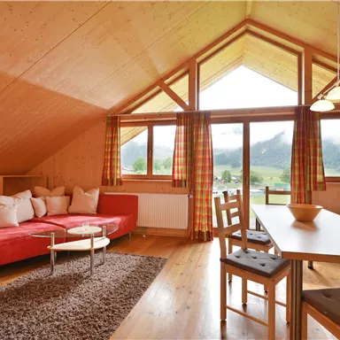 Ein gemütliches Wohnzimmer mit einem roten Sofa und einem Esstisch. Große Fenster bieten einen schönen Blick auf die Berge und lassen viel Licht herein.