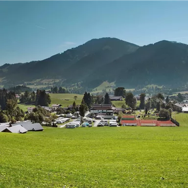 Eine idyllische Landschaft mit grünen Wiesen und Bergen im Hintergrund. Im Vordergrund sind einige Gebäude und Tennisplätze zu sehen.