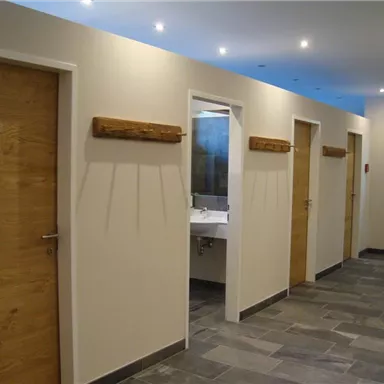 Ein moderner Flur mit mehreren Holztüren und einem Waschbereich. Der Boden ist mit grauen Fliesen ausgelegt, und die Wände sind hell gestrichen.