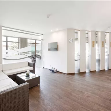 Ein modernes Wohnzimmer mit einem hellen, offenen Raum. Es gibt eine weiße Couch, Holzfußboden und große Fenster, die viel Licht hineinlassen.