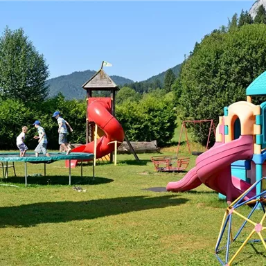 A colorful playground with a slide, a trampoline, and a play tower. Two children are happily jumping on the trampoline in the green garden.