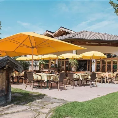 Ein einladendes Restaurant mit gelben Sonnenschirmen und gemütlichen Sitzgelegenheiten im Freien. Umgeben von Grünflächen und Bäumen bietet es eine entspannte Atmosphäre.