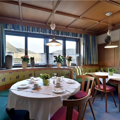 Ein gemütliches Esszimmer mit einem runden Tisch und Sitzbänken. Durch die Fenster kann man die umliegende Landschaft sehen.