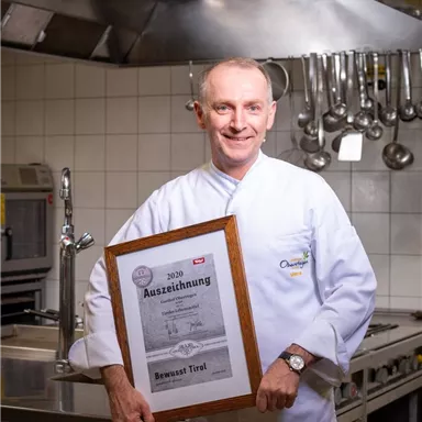Ein Koch in einer professionellen Küche hält stolz eine Auszeichnung in der Hand. Im Hintergrund sind Küchengeräte und Töpfe sichtbar.
