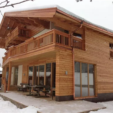 Ein modernes Holzhaus im Schnee mit großen Fenstern und einem Balkon. Die Umgebung ist verschneit und vermittelt eine gemütliche Atmosphäre.