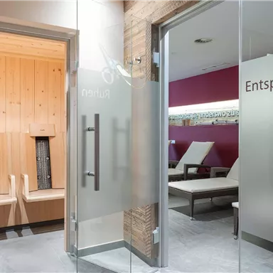 A bright wellness area with a sauna in the background and lounge chairs. The glass doors are labeled with the words "Relax" and "Enjoy."