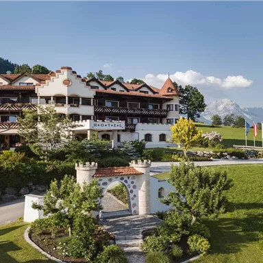 A charming hotel with traditional architecture, surrounded by green meadows and trees. In the background, mountains and a clear sky are visible.