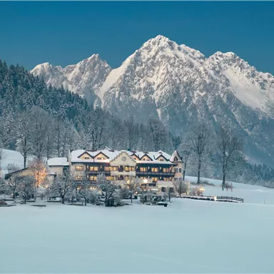 A picturesque winter landscape with snow-covered mountains in the background. In the foreground stands a cozy hotel, surrounded by snow-covered trees.