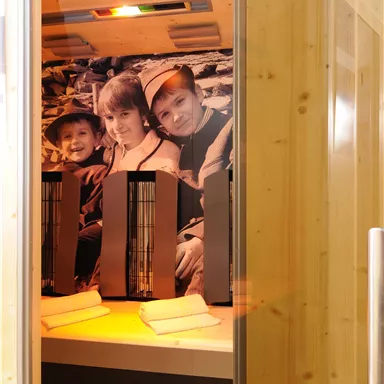 Ein Innenraum einer Sauna mit Holzverkleidung und einem Bild von drei lächelnden Kindern an der Wand. Im Vordergrund stehen zwei Heizstrahler auf einer Bank.