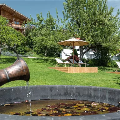 A beautiful garden with a pond and a water jug in the foreground. In the background, there are loungers and sun umbrellas under a tree.