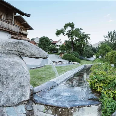 A quiet garden with a stone water feature. In the background, there are wooden houses and green trees.