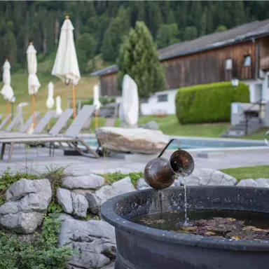 A quiet outdoor area with a swimming pool, loungers, and sun umbrellas. In the foreground, water flows from a decorative bowl.