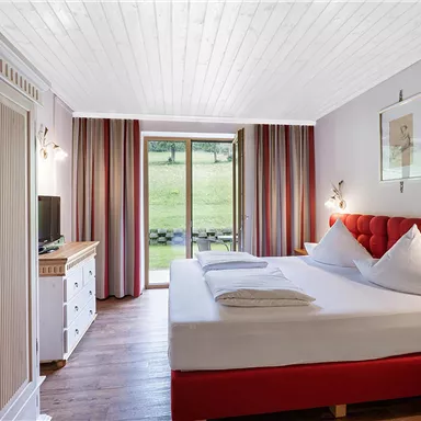 A modern bedroom with a red bed and two pillows. Large windows offer a view of the green landscape.