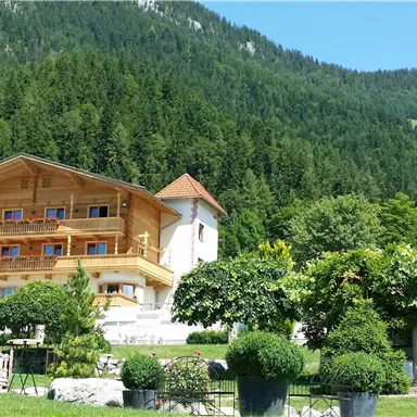 A cozy wooden house amidst a green mountain landscape. The sky is clear and the surroundings are lushly overgrown.