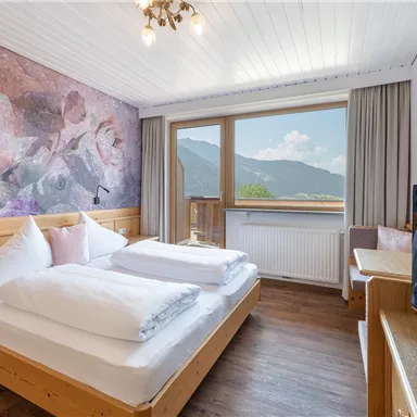 A cozy hotel room with two beds and wooden decor. Through the window, one looks out onto a picturesque mountain landscape.