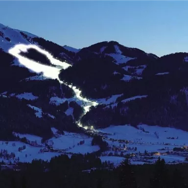 Eine schneebedeckte Berglandschaft bei Nacht mit beleuchteten Pisten. Im Hintergrund sind dunkle Berge und ein klarer Himmel zu sehen.