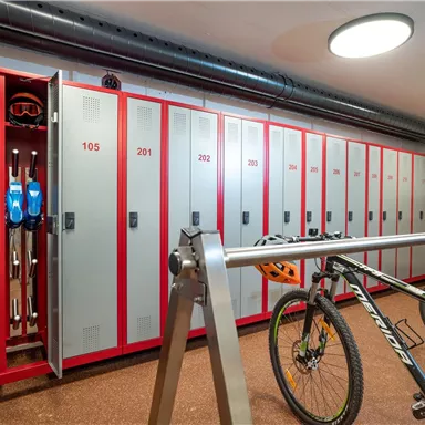 Ein geräumiger Schrankraum mit vielen Schließfächern. Ein Fahrrad steht neben einem offenen Schließfach, in dem Skiausrüstung aufbewahrt ist.